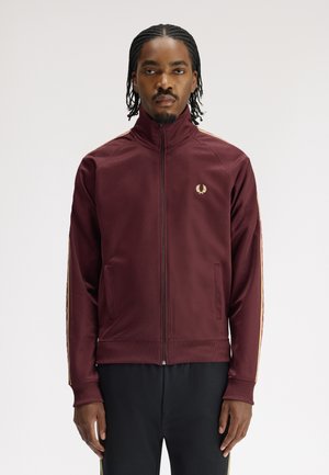 Mann mit geflochtenen Haaren, der eine bordeauxrote Trainingsjacke mit Reißverschluss, Goldstreifen und Emblem trägt, vor einem schlichten weißen Hintergrund steht.