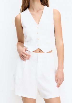 Femme portant une veste blanche sans manches boutonnée et un short blanc taille haute, debout avec une main posée sur sa taille.