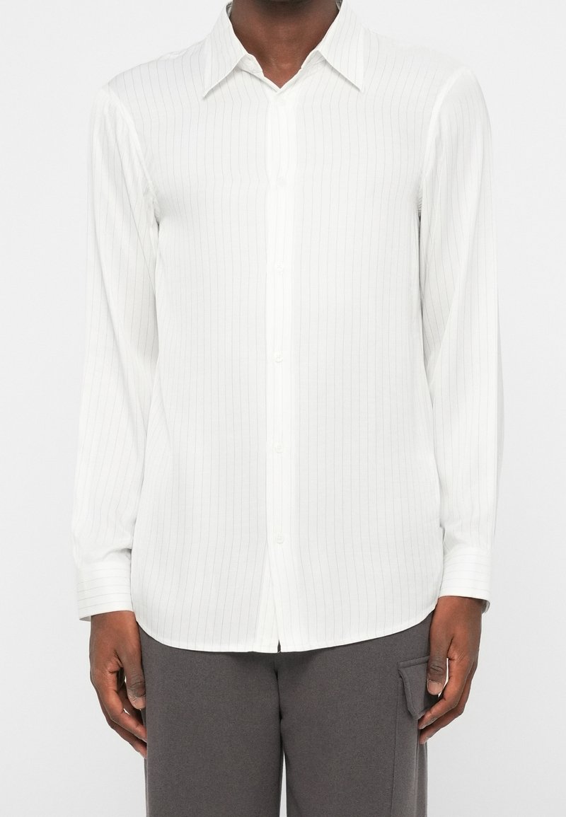Homme portant une chemise blanche à manches longues avec de fines rayures verticales, associée à un pantalon gris foncé avec une poche latérale.