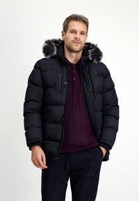 Giubbotto a sacco nero con cappuccio foderato in pelliccia, imbottito con isolamento. L'uomo indossa un maglione di lana viola sotto, con un motivo testurizzato.