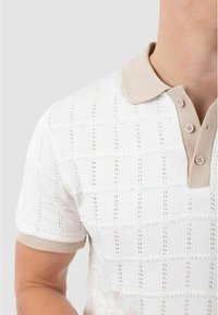 Polo en tricot blanc avec un motif texturé, col beige contrastant et patte de boutonnage, manches courtes avec un bord beige assorti.