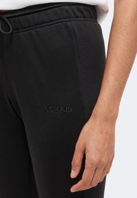 Άτομο με μαύρο παντελόνι Adidas με ελαστική μέση και κορδόνι, το δεξί χέρι φαίνεται χαλαρό δίπλα στο σώμα.