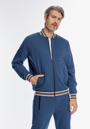 Mężczyzna w wieku średnim z siwymi włosami, ubrany w niebieską kurtkę na zamek i pasujące spodnie z paskowanymi mankietami i kieszeniami, pozujący na białym tle.