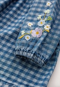 Blue gingham fabric with embroidered floral accents in yellow, purple, and white. Features gathered elastic cuffs along the sleeves.