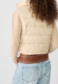 Beżowa pikowana kamizelka z dzianinowymi rękawami, przewężona w talii brązowym paskiem z metalowymi ćwiekami, w połączeniu z jasnoniebieskimi dżinsami.