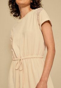 Robe beige avec des manches courtes retroussées, des accents en dentelle, une taille élastique et un détail de cordon noué. Texture de tissu lisse et douce.