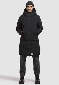 Sort polstret vinterjakke med hætte, der har en frontlynlås, sidelommer og ribbede manchetter. Båret med mørke jeans og sorte snørebodssko.