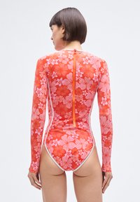 Červené plavky s dlouhým rukávem s květinovým vzorem a zipem na zádech. Mají vzor růžových květin a kontrastní bílé lemování.