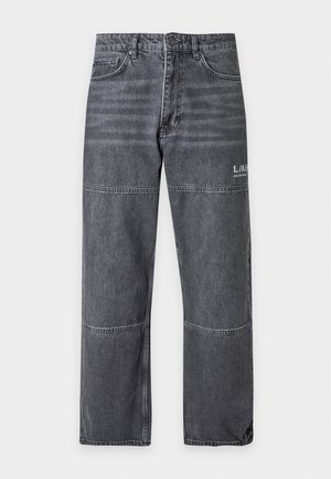 Jeune homme portant un t-shirt blanc à motif, un jean gris ample avec un texte blanc, et des baskets noires et blanches, debout avec une main dans la poche.