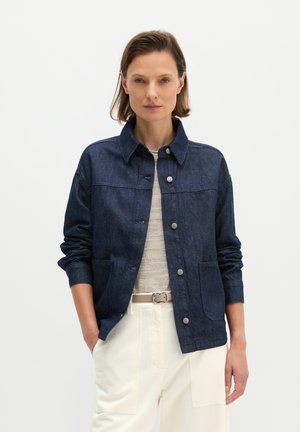 Frau mit kurzen braunen Haaren, die eine dunkelblaue Jeansjacke, einen hellbeigen Pullover, einen hellbeigen Gürtel und weiße Hosen trägt, steht mit einer Hand in der Tasche.