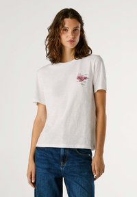 T-shirt en coton blanc à manches courtes, avec un petit design graphique rose sur la poitrine. Coupe décontractée, texture lisse.