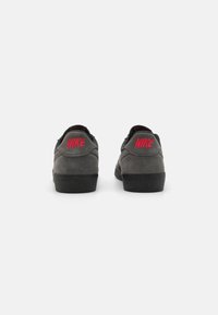 Sive semiš tene s črnimi gumijastimi podplati, ki imajo na peti rdečo vezeno Nike oznako. Nizka zasnova z gladko teksturo.