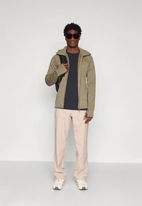 Olijfgroene jas met ritssluiting, donkerblauw shirt, lichtbeige broek en witte sneakers. De persoon draagt een zwarte rugzak en een zonnebril.