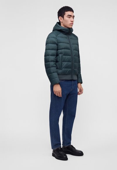Homme portant une veste matelassée verte avec capuche, un jean bleu et des chaussures habillées noires, debout devant un fond blanc uni.