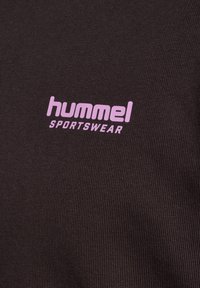 Krupni plan crne tkanine s ružičastim logotipom "hummel SPORTSWEAR" otisnutim u sredini, prikazujući teksturirani pleteni materijal.