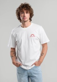 White cotton t-shirt featuring a red sun graphic with the text "SUNSET BOULEVARD." Short sleeves and a crew neckline. Light blue jeans.