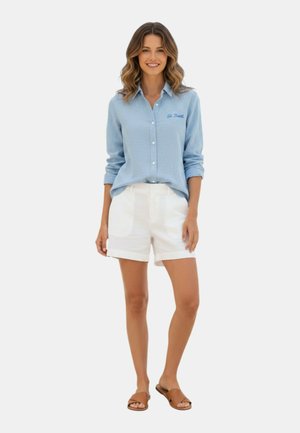 Donna sorridente che indossa una camicia azzurro chiaro con bottoni con testo "St. Barth", pantaloncini bianchi e sandali marroni, in piedi su uno sfondo semplice.