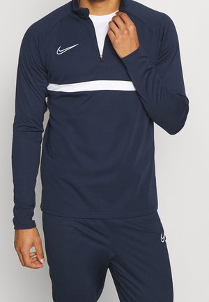 Mężczyzna ubrany w granatową sportową bluzę Nike z białym poziomym paskiem i dopasowane spodnie, regulujący zamek przy kołnierzu.