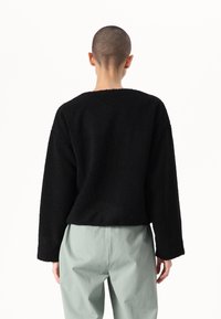 Maglione nero, strutturato, con scollatura rotonda ampia e maniche lunghe. Indossato con pantaloni verde chiaro, che mostrano una vestibilità ampia e rilassata.