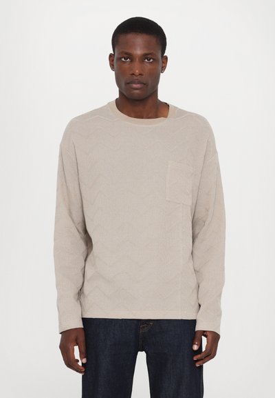 Maglione beige a maniche lunghe con un motivo a zig zag testurizzato, scollo rotondo e taschino sul petto sinistro. Abbinato a jeans scuri.