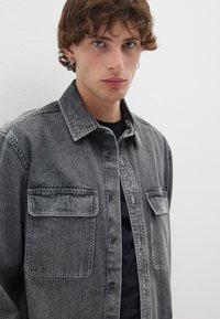 Camicia in denim grigia con colletto, chiusura a bottoni e due tasche sul petto. Presenta una texture leggermente sbiadita e un design con vestibilità rilassata.