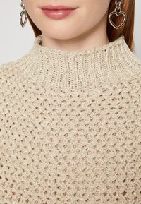 Gros plan d'une personne portant un pull en maille beige avec un col côtelé et des boucles d'oreilles argentées en forme de cœur.