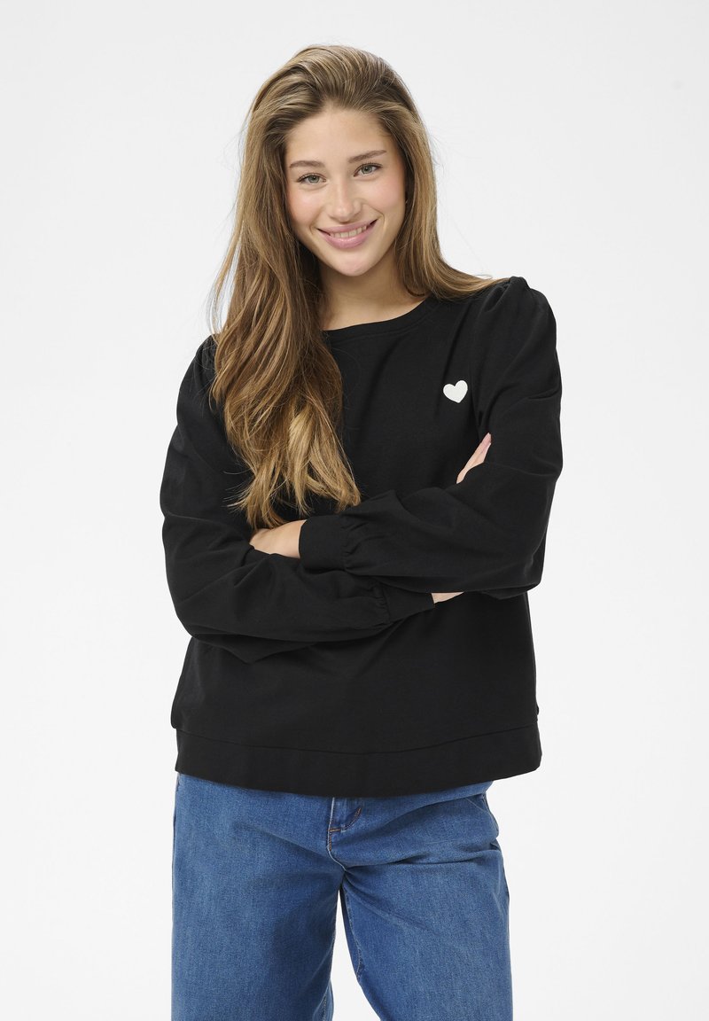 Schwarzer Pullover mit langen, gepufften Ärmeln und rundem Ausschnitt; mit einem kleinen weißen Herzemblem auf der Brust. Kombiniert mit blauen Jeans.