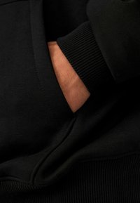 Sort sweatshirt med ribbede mansjetter og en hånd i en forlommes, der viser den glatte stoftekstur og det sømløse design.
