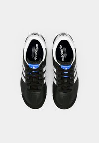 Fekete Adidas cipőpár fehér csíkokkal, kék logócímkékkel és fekete fűzőkkel, felülről szürke háttéren.