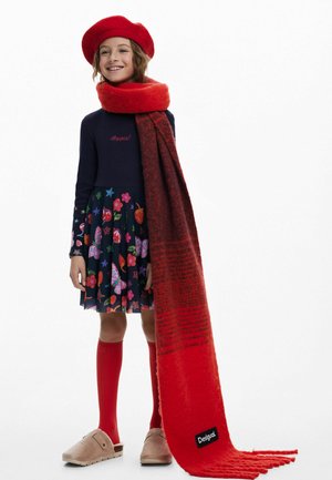 Rote Mütze, langer roter Schal mit Farbverlauf, schwarze Markenkennzeichnung, marineblaues Kleid mit buntem Blumen- und Schmetterlingsmuster, rote Kniestrümpfe, beigefarbene Clogs.