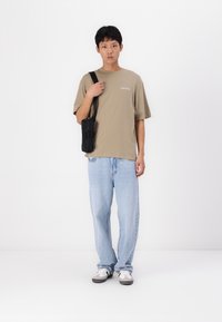 Camiseta beige de gran tamaño con texto en la parte frontal, jeans holgados azules claros y bolso cruzado negro; complementada con zapatillas grises y blancas.