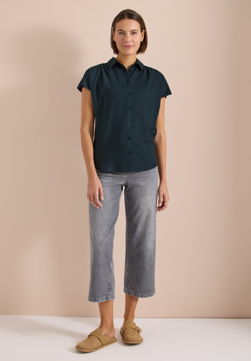 Femme debout contre un mur beige, vêtue d'une chemise foncée à manches courtes boutonnée, d'un jean gris court et de chaussures beige à enfiler, souriant doucement.
