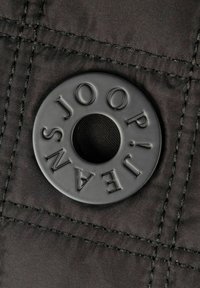 Czarna tkanina z pikowanym ściegiem zawiera okrągły czarny plastikowy oczko z wytłoczonym napisem "JOOP!" i "JEANS", z centralnym otworem.