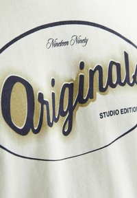 Witte t-shirt van stof met "Original" in donkerblauwe cursieve letters, met een gradient gouden achtergrond, en "Nineteen Ninety" erboven in kleinere letters.