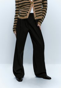 Pantalons noirs à jambe large avec une texture lisse ; associés à un cardigan rayé brun et noir, avec des boutons et une coupe décontractée.