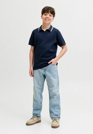 Junge lächelt, steht mit einer Hand in der Tasche, trägt ein dunkelblaues Polohemd mit kurzen Ärmeln, hellblaue Jeans und beige Schuhe vor schlichter Hintergrund.