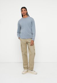 Šviesiai mėlyna ilgasleevių marškiniai, šviesiai rudi šoninėmis kišenėmis cargo kelnės ir baltos sportbačiai, stovintys prieš paprastą baltą foną.