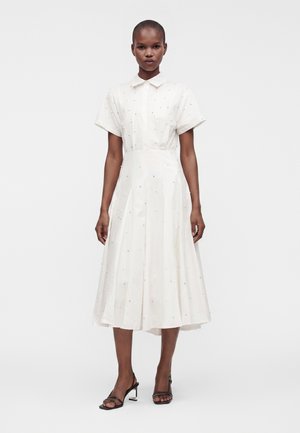 Femme portant une robe midi blanche à manches courtes, col avec une coupe en ligne A, ornée de motifs subtils à pois, et des sandales à talons noires à bout ouvert, debout.