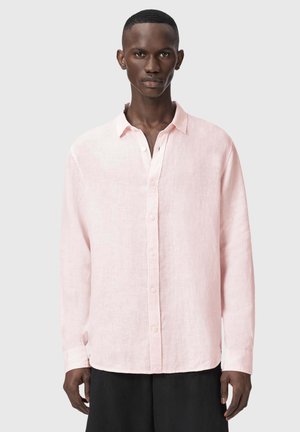 Mand iført en lyserød langærmet skjorte med knapper og sorte bukser, stående foran en ensfarvet lysegrå baggrund.