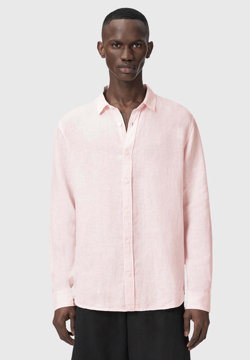 Uomo che indossa una camicia rosa chiaro a maniche lunghe con bottoni e pantaloni neri, in piedi davanti a uno sfondo grigio chiaro uniforme.