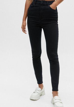 Personne portant un jean skinny noir taille haute et des mocassins blancs épais, une main dans la poche, sur un fond uni.