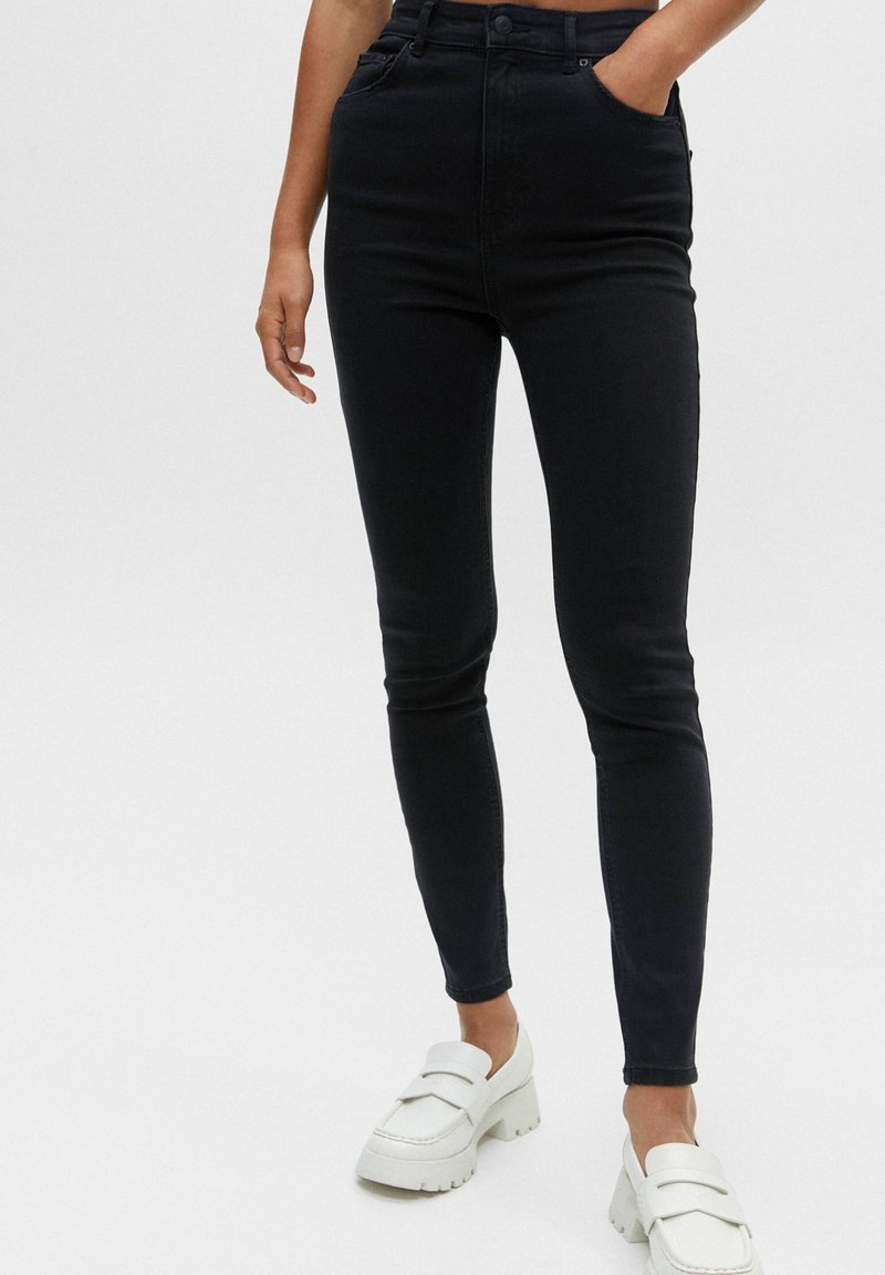 Personne portant un jean skinny noir taille haute et des mocassins blancs épais, une main dans la poche, sur un fond uni.