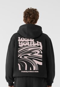 Crna dukserica s velikim ružičastim i bijelim spiralama na leđima s natpisima "izgubljena mladost" i "Kreativnost se nalazi u kaosu."