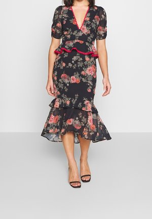 Kobieta ubrana w czarną sukienkę w kwiaty z czerwonym obszyciem, z paskiem w talii typu peplum, falbanianym dołem i krótkimi rękawami, zestawioną z czarnymi sandałami na otwartym palcu.