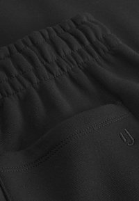 Zwarte kledingstuk met een gestructureerde tailleband en een vlakke zak. Voorzien van subtiele stiksels en een klein logodetail. Glad, zacht materiaal.