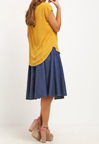 Haut jaune à manches courtes avec un ourlet arrondi, associé à une jupe en denim bleu foncé, de design évasé et de coupe mi-genou. Sandales à talons hauts rose clair.