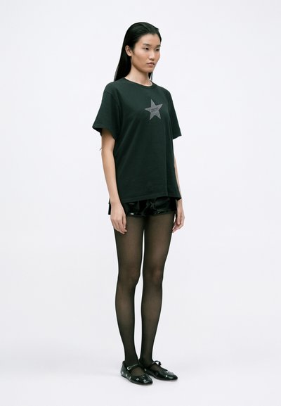 Tricou din bumbac negru cu un grafic de stea argintie, combinat cu pantaloni scurți negri lucioși și dresuri negre transparente, completat cu pantofi negri fără toc.
