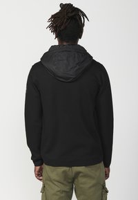 Czarny sweatshirt z teksturowanym kapturem, o gładkim wykończeniu. Noszony z oliwkowymi spodniami cargo, prezentującymi swobodny design i fason.