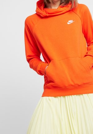 Osoba ubrana w pomarańczową bluzę Nike z kapturem, z rękami w przedniej kieszeni, oraz w jasnżółtą plisowaną spódnicę na jednolitym tle.