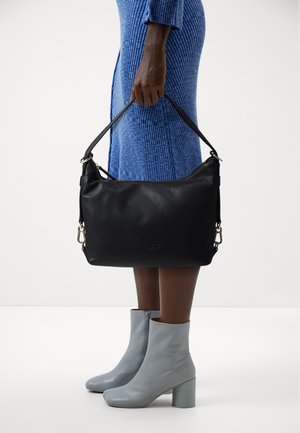 Sac à bandoulière en cuir noir à forme arrondie, orné de détails en métal doré et d'une surface texturée, porté par une personne vêtue d'une robe bleue côtelée.